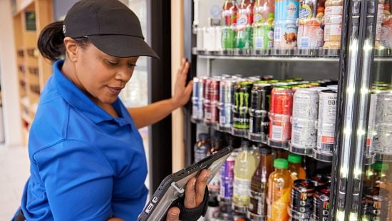 Worker in blue uniform using handheld device to restock beverages in refrigerated display case.