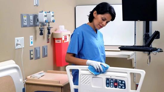Healthcare staff member wearing gloves cleaning a hospital bed control panel with a disinfectant wipe in a patient room.